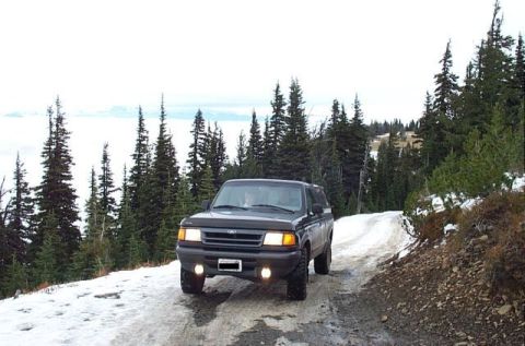 1995 Ford Bronco - A dark SUV navigates a snowy mountain road surrounded by pine trees, likely a 1995 Ford Explorer.