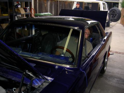 1965 Ford Mustang - A person sits in a customized coupe with the hood open, while a white truck is parked behind it.