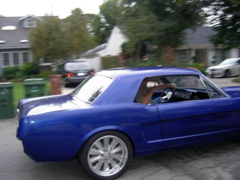 1965 Ford Mustang - A blue 1965 Ford Mustang coupe is seen driving on a street with houses in the background.