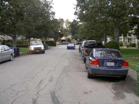1965 Ford Mustang - A blue coupe is seen driving away on a street lined with parked cars and trees.