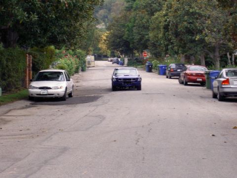 1965 Ford Mustang - A blue classic car is seen driving down a tree-lined street with other vehicles parked along the side.