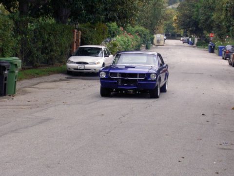 1965 Ford Mustang - A blue 1965 Ford Mustang coupe is seen driving down a residential street with a white sedan parked nearby.