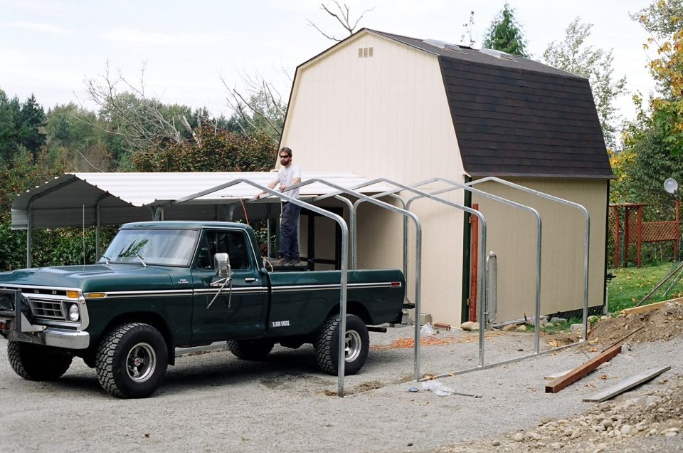Dark green classic Ford pickup by a carport frame under construction, man standing in the bed, barn-style shed behind