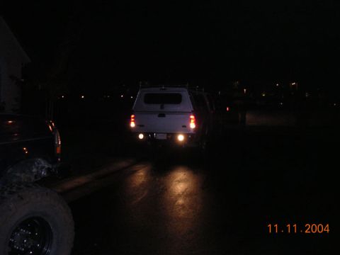 2004 Ford F-250 - A white pickup truck with a camper shell is seen from the rear at night, with its reverse lights on.