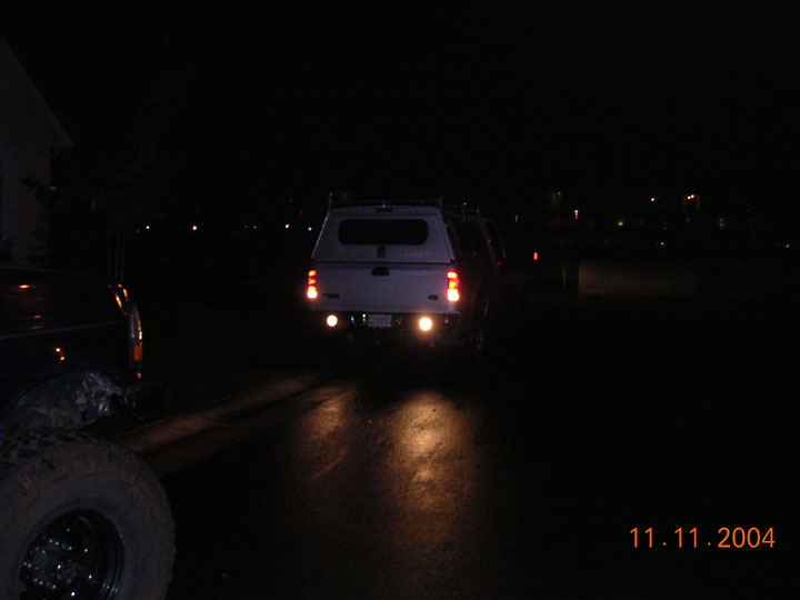 White pickup with camper shell at night, rear bumper flood lights lighting the pavement