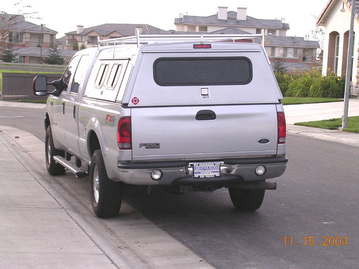 Silver Ford F-250 FX4 with camper shell and bumper flood lights, rear view at curb