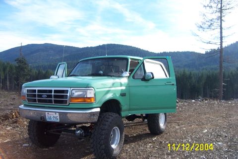 1996 Ford Bronco - This lifted green Ford Bronco is parked outdoors with its doors open, set against a backdrop of forested hills.