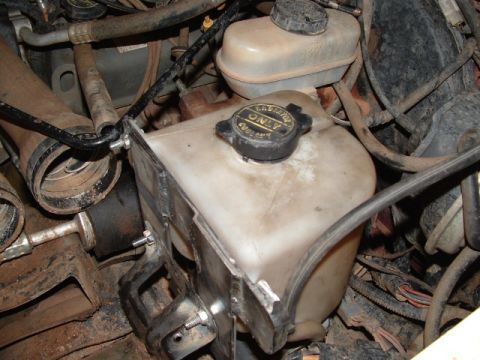 1990 Ford Bronco - Grimy engine-bay close-up of a translucent plastic coolant degas/overflow bottle in a metal bracket, its black cap embossed with a "Lubrica…