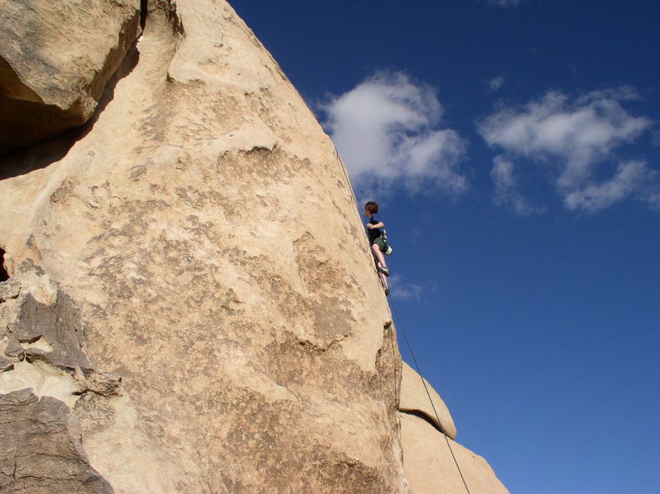 Rock climber ascends a large boulder under a blue sky with scattered clouds. Watermark reads SUPERMOTORS.NET