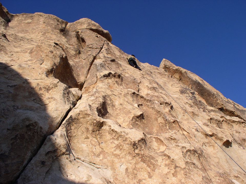 Rock climber ascending a large rock face under a clear blue sky. Rope visible