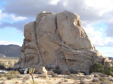 1990 Ford Bronco - Climbers ascend a massive rock formation in a desert landscape, with vehicles visible at the base.