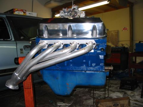 1985 Ford Crown Victoria LTD - A blue V8 engine with chrome valve covers and headers is displayed, with a light blue car in the background.
