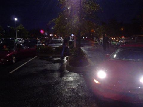 1996 Ford Probe - A red sports car is parked in a lot at night, with its headlights illuminated. Several other vehicles and people are present.