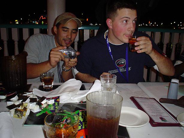 Two men sit at a table with drinks, one taking a photo with a digital camera