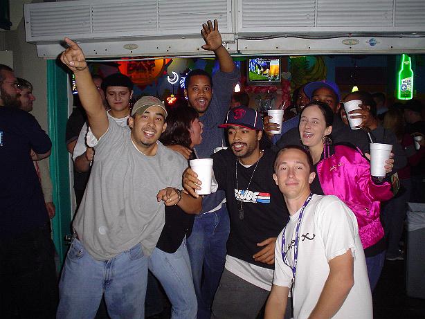 Group of smiling people holding drinks in a bar, one points up