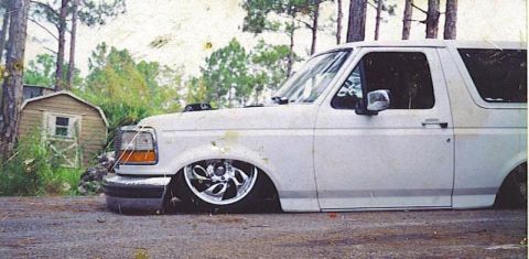 1985 Ford Bronco - A white full-size Ford Bronco slammed low to the ground, shown from a 3/4 front angle with body panels nearly touching the pavement and chr…