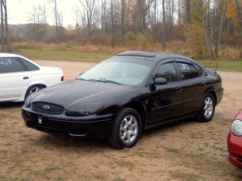 2004 Ford Taurus - A black sedan is parked on a dirt lot with trees in the background. Another car is visible to the left and a portion of a red car is on the…