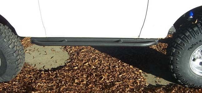 Black tubular rock-slider rocker bar mounted between two mud tires over wood chips