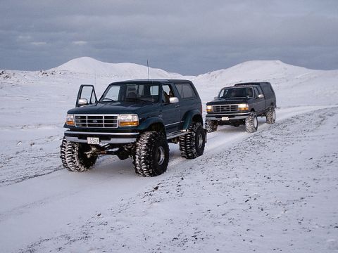1996 Ford Bronco - Two modified Ford Broncos navigate a snowy landscape under a cloudy sky.