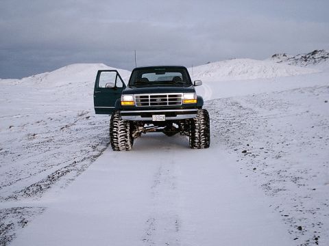 1996 Ford Bronco - A modified Ford pickup truck with its door open sits on a snow-covered road in a desolate, hilly landscape.