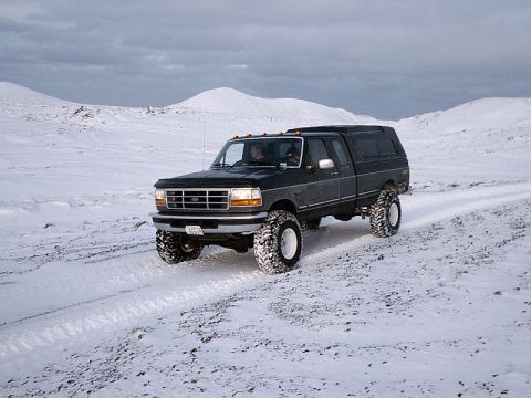 1996 Ford Bronco - A lifted black pickup truck with a camper shell navigates a snowy landscape, its large tires kicking up snow.