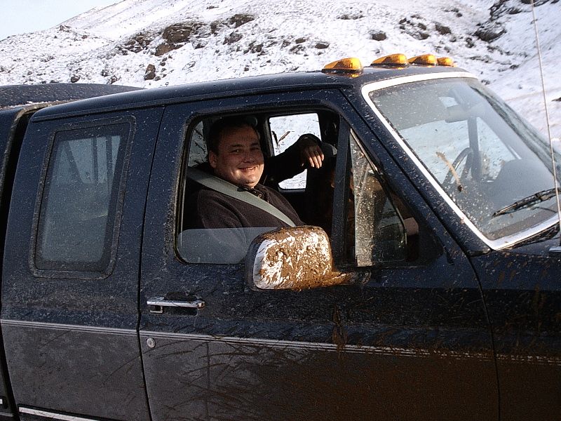 Man smiling in cab of mud-splattered dark blue Ford Bronco on snowy hill