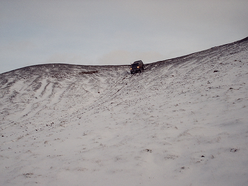 Dark SUV with headlights on driving down a snowy hill under gray sky