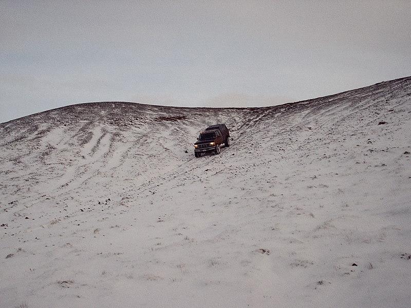 Dark SUV with headlights on climbing a snowy hill under gray sky