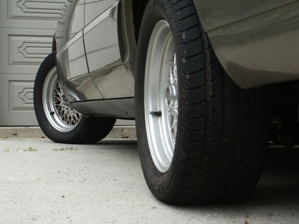 Low-angle view of gray car's right-side silver wire wheels with Dunlop tires on driveway.