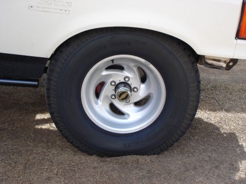 1987 Ford Bronco - A close-up view of a vehicle's wheel and fender, showing aftermarket rims and a portion of the body.