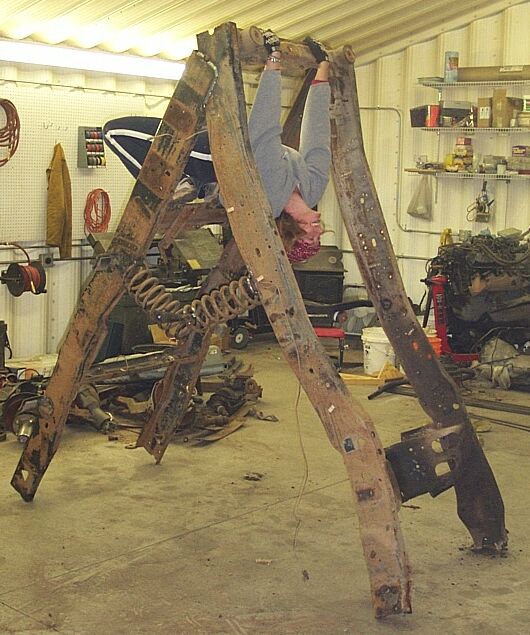 Bare rusty truck frame stood upright in a shop with person hanging from it