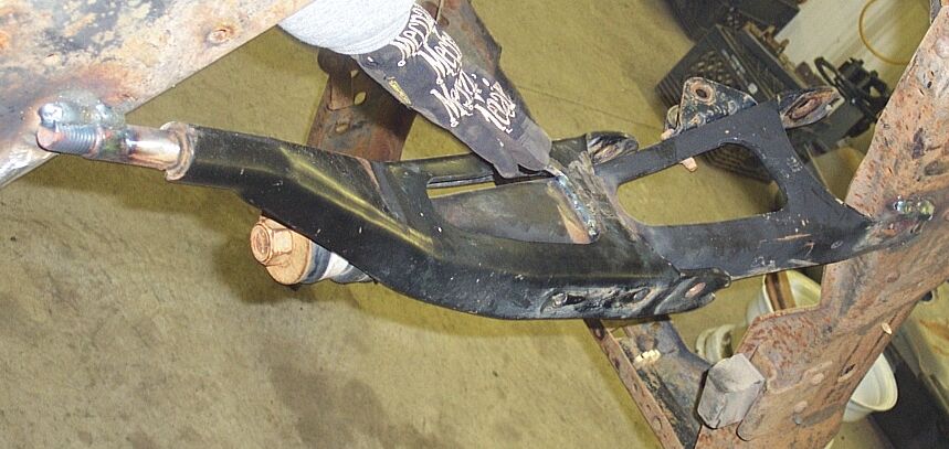 Gloved hand holding a black radius arm test-fitted between rusty frame rails