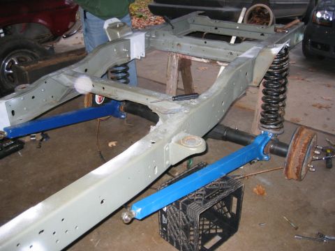 1978 Ford Bronco - A bare truck frame painted light gray sitting on jack stands in a garage during a suspension rebuild, with blue-painted radius arms and a c…