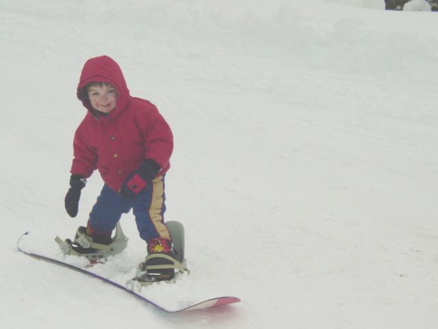 Small child in red coat and blue pants riding a snowboard down a snowy slope