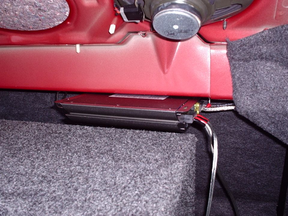 Red car interior panel with a black amplifier mounted below a speaker. Carpeted surfaces are visible