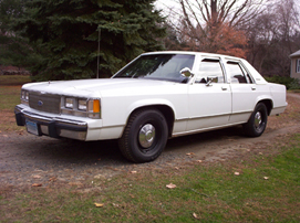1991 Ford LTD Crown Victoria - A white boxy Ford full-size four-door sedan parked on a gravel driveway with bare trees and a lawn behind it, seen from the front three-qua…