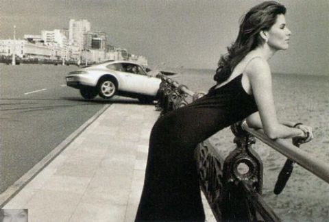 1990 Ford Bronco - A woman observes a Porsche 911 as it drives past a coastal city.