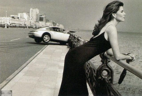 Woman in black dress leans on railing, looking out at sea. Silver Porsche drives on road behind her. "SUPERMOTORS www.supermotors.net" logo visible