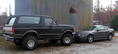 1988 Ford Bronco - A black Ford Bronco is parked next to a gray Ford Mustang in front of a corrugated metal building.