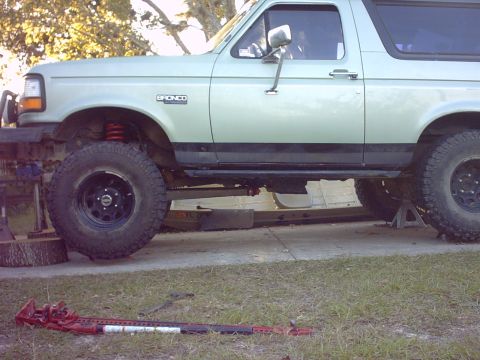 1995 Ford Bronco - Driver-side view of a light green 1990s Ford Bronco lifted with red coil springs and mud-terrain tires on black steel wheels, its rear axle…