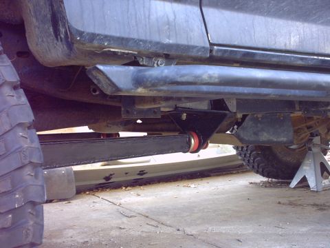 1995 Ford Bronco - Low ground-level view under the driver's side of a lifted truck showing a fabricated black rocker-guard bar bolted to a frame bracket, with…