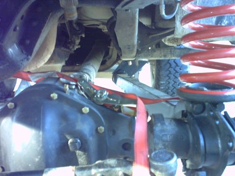 1995 Ford Bronco - Close underbody shot of a lifted 4x4 with a red-painted coil spring, showing the transfer case housing, driveshaft with U-joint, leaf sprin…