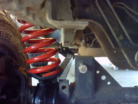 1995 Ford Bronco - Undercarriage view of a lifted truck showing a bright red front coil spring seated on a black axle bracket, with an all-terrain tire at lef…