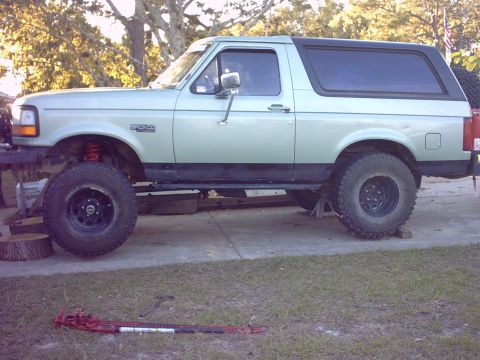 1995 Ford Bronco - Driver-side profile of a light seafoam-green 1992–96-style Ford Bronco with a dark rear hardtop, lifted on red coil springs and tall all-te…