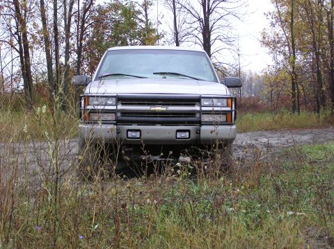 2000 Chevrolet Tahoe - This silver SUV, likely from the mid-1990s, is seen from the front, partially obscured by tall grass and mud.