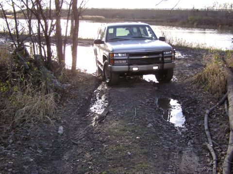 2000 Chevrolet Tahoe - A silver SUV navigates a muddy path beside a body of water.