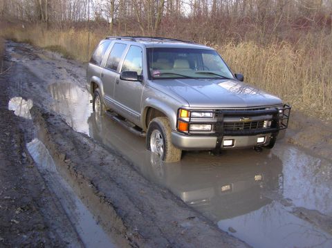 2000 Chevrolet Tahoe - Silver SUV with a grille guard is stuck in a muddy road, tires submerged.