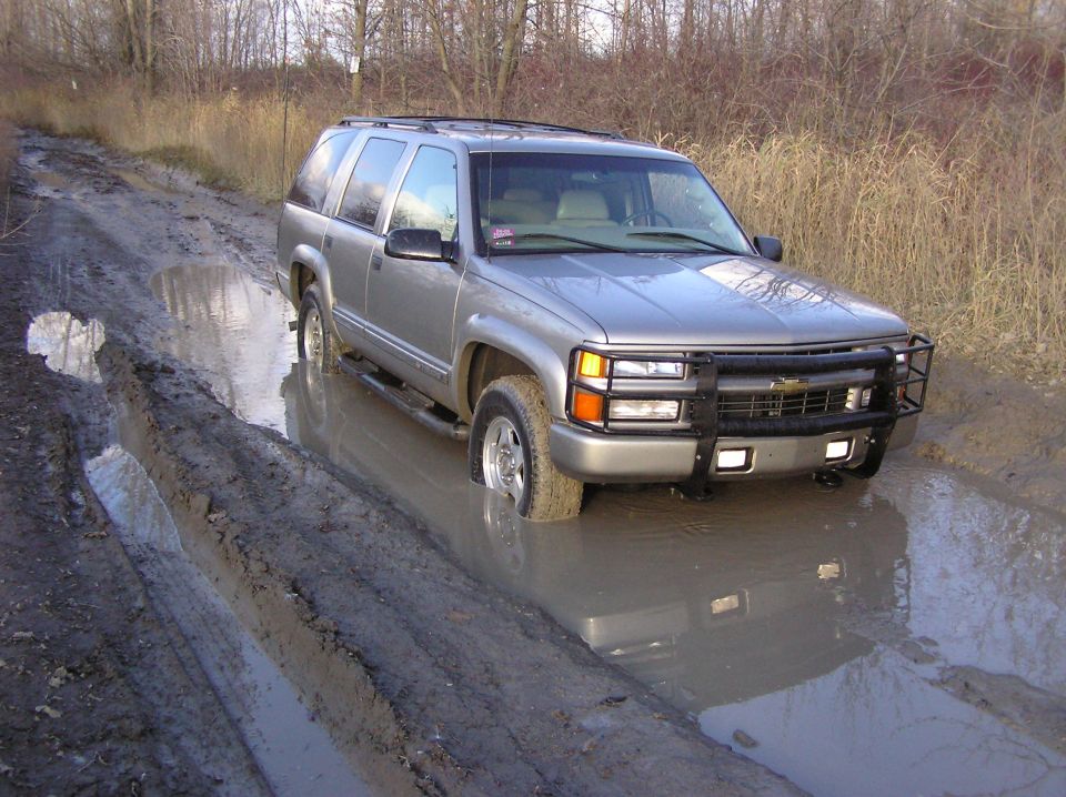 Silver SUV stuck in a muddy rut on a dirt road. Front grille guard is visible