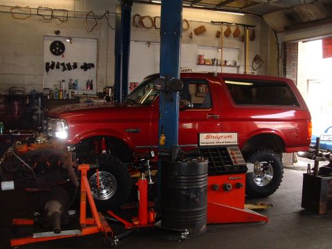 1994 Ford Bronco - Red lifted Ford Bronco with hardtop and headlights on, parked driver's-side-on inside a working repair shop between a two-post lift column…