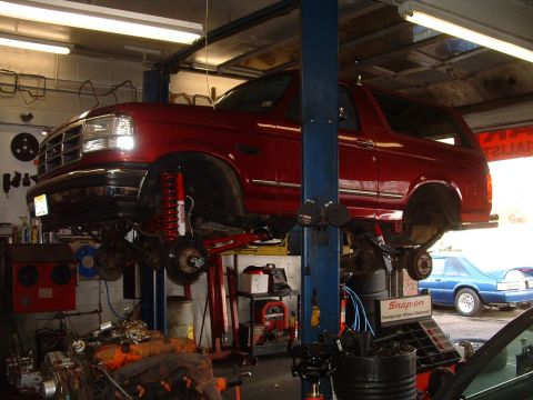 1994 Ford Bronco - Red 1994 Ford Bronco undergoing service on a lift in a repair shop, with a blue car in the background.